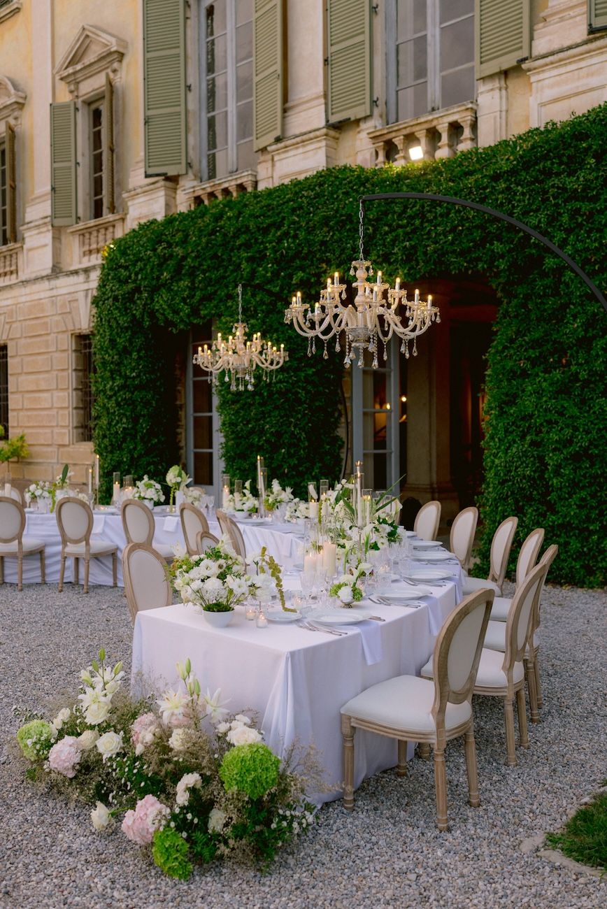 Wedding dinner setup with white, pale pink and green flowers on semi circular tables enlightened by glass chandeliers. Design Solidea Events