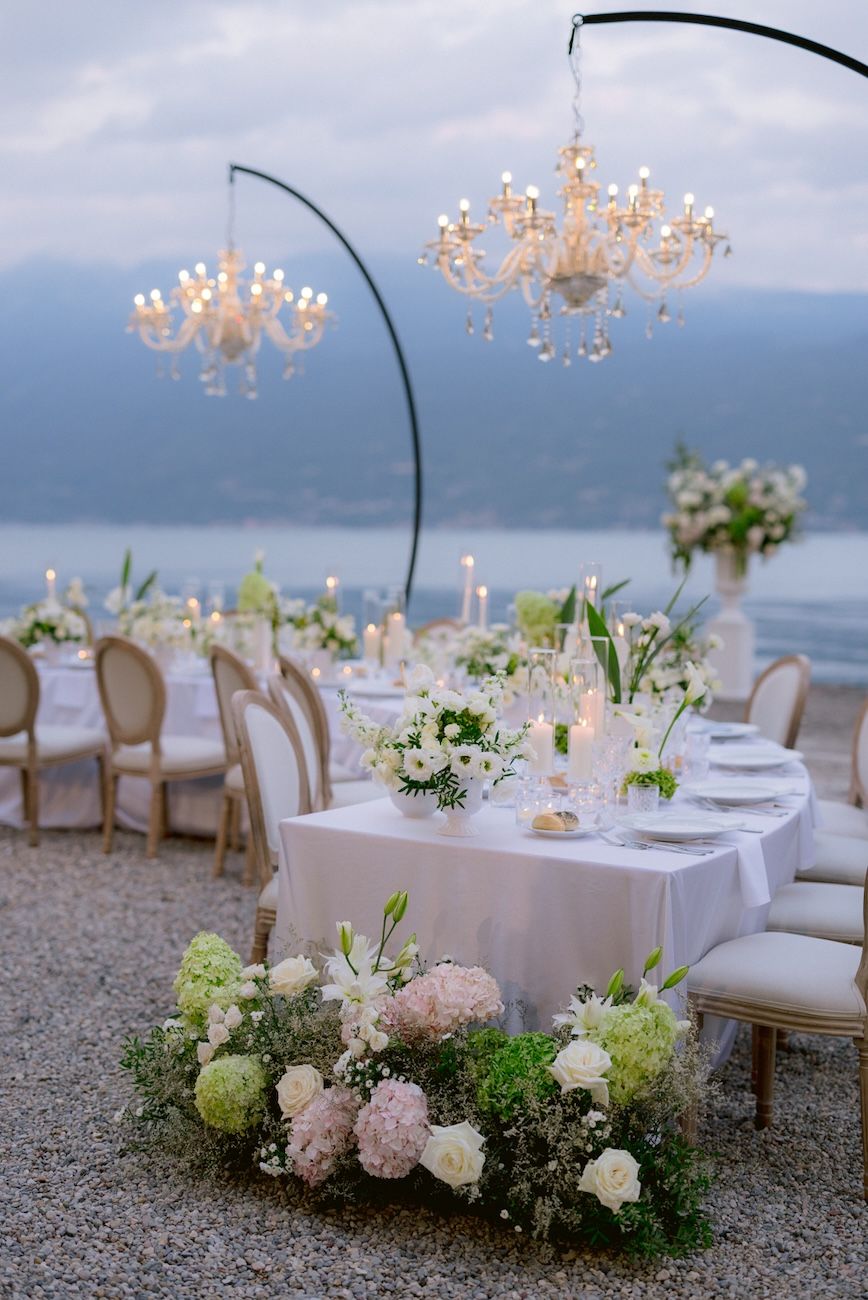 Wedding dinner setup with white, pale pink and green flowers on semi circular tables enlightened by glass chandeliers. Design Solidea Events