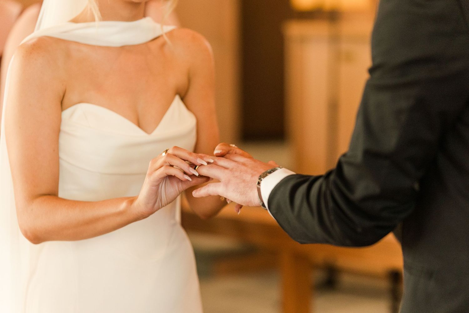 Bride in white strapless gown placing wedding ring on groom's finger during ceremony.