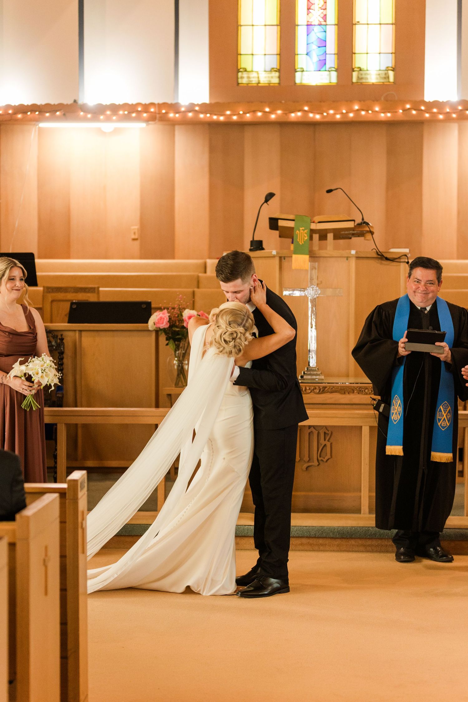 Bride and groom share first kiss at church altar while officiant smiles and bridesmaid watches during wedding ceremony.
