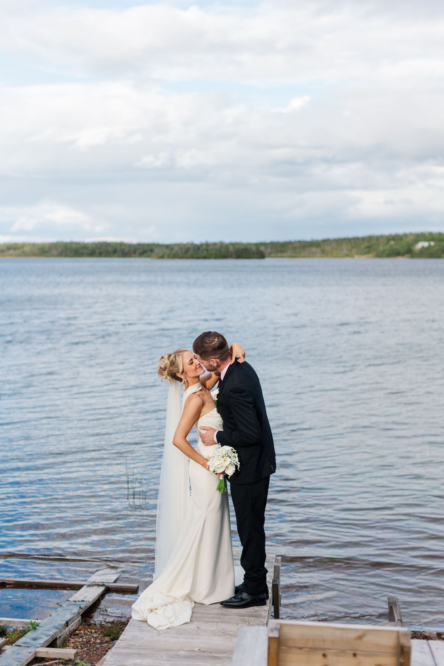 A bride and groom share a kiss on a wooden dock by a serene lake on their wedding day.