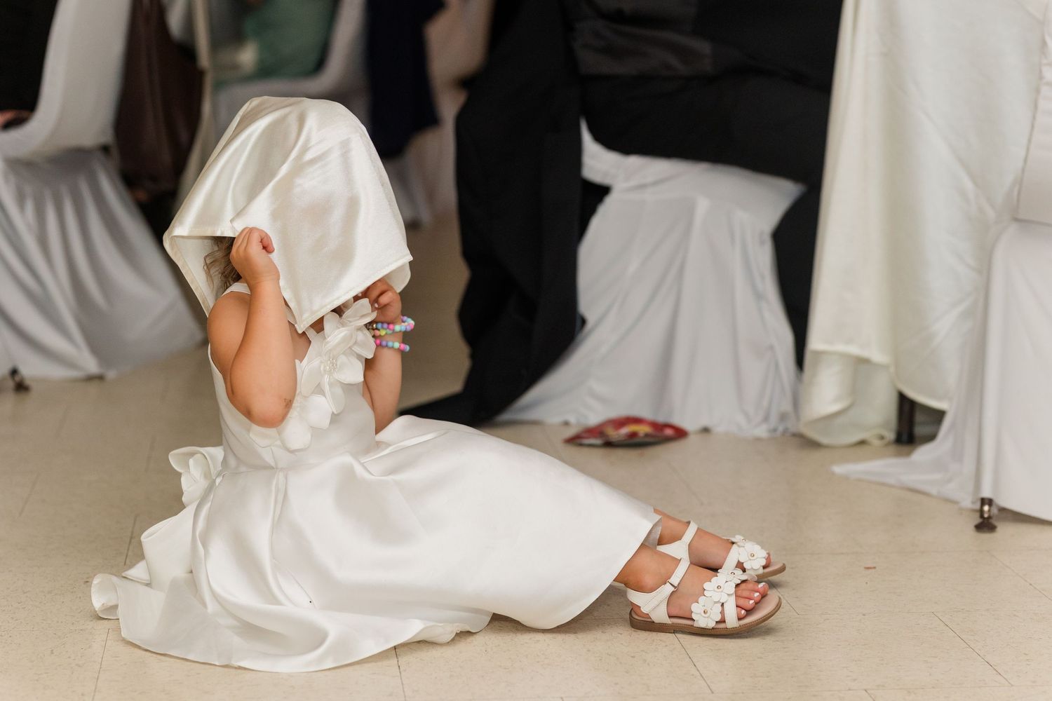 A young flower girl in a white dress sits on the floor pulling her veil over her face at a wedding reception.