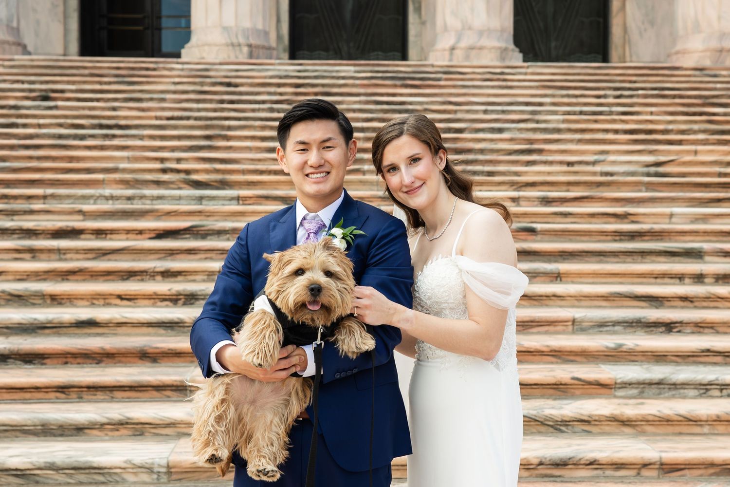 A smiling couple in wedding attire holds a small brown dog on stone steps.