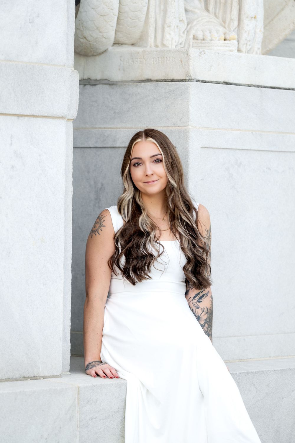 Young woman with long wavy hair and tattoos poses in white dress against marble columns.