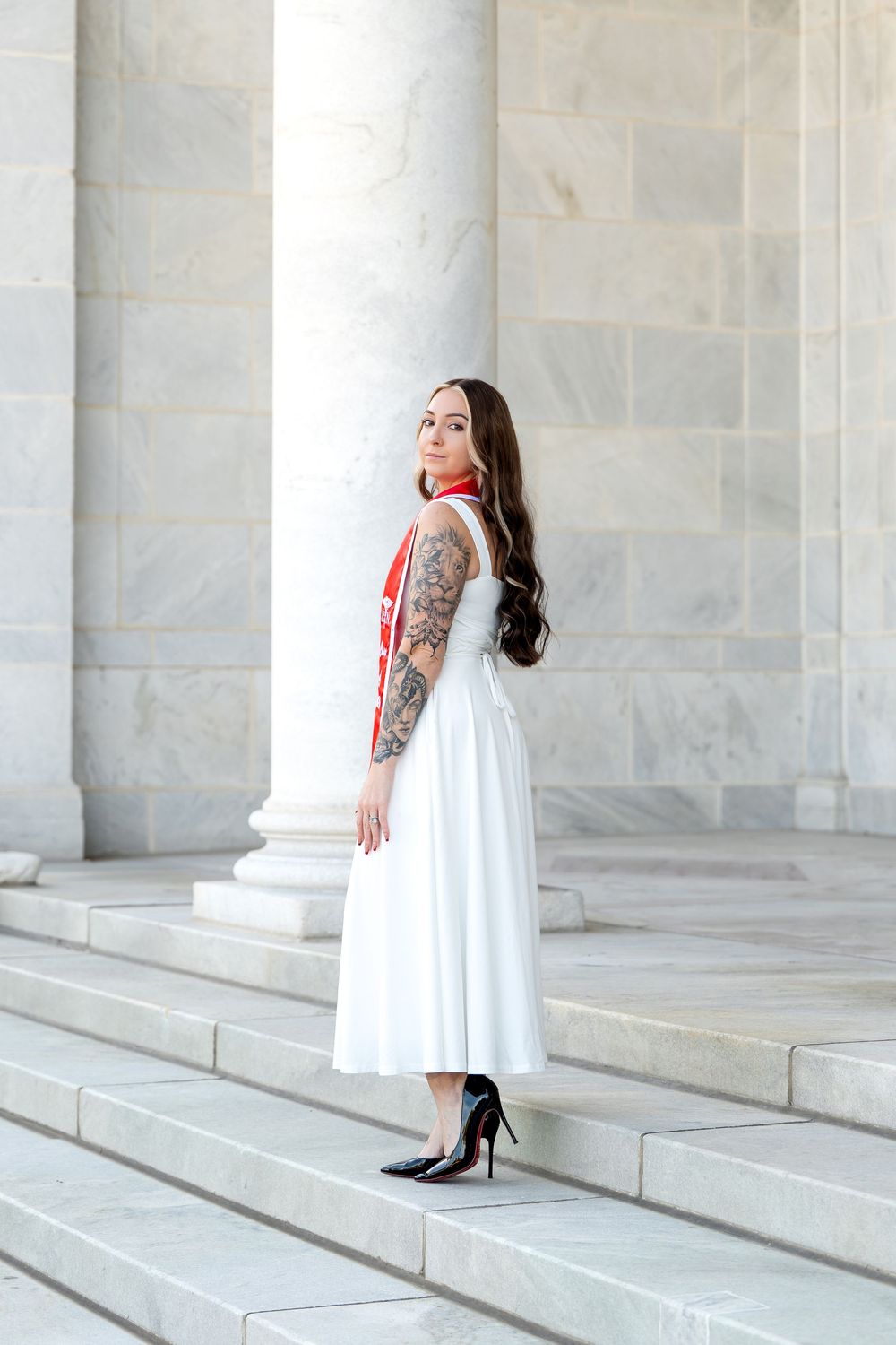 Young woman with sleeve tattoo in white midi dress and heels poses on marble steps with red graduation sash.