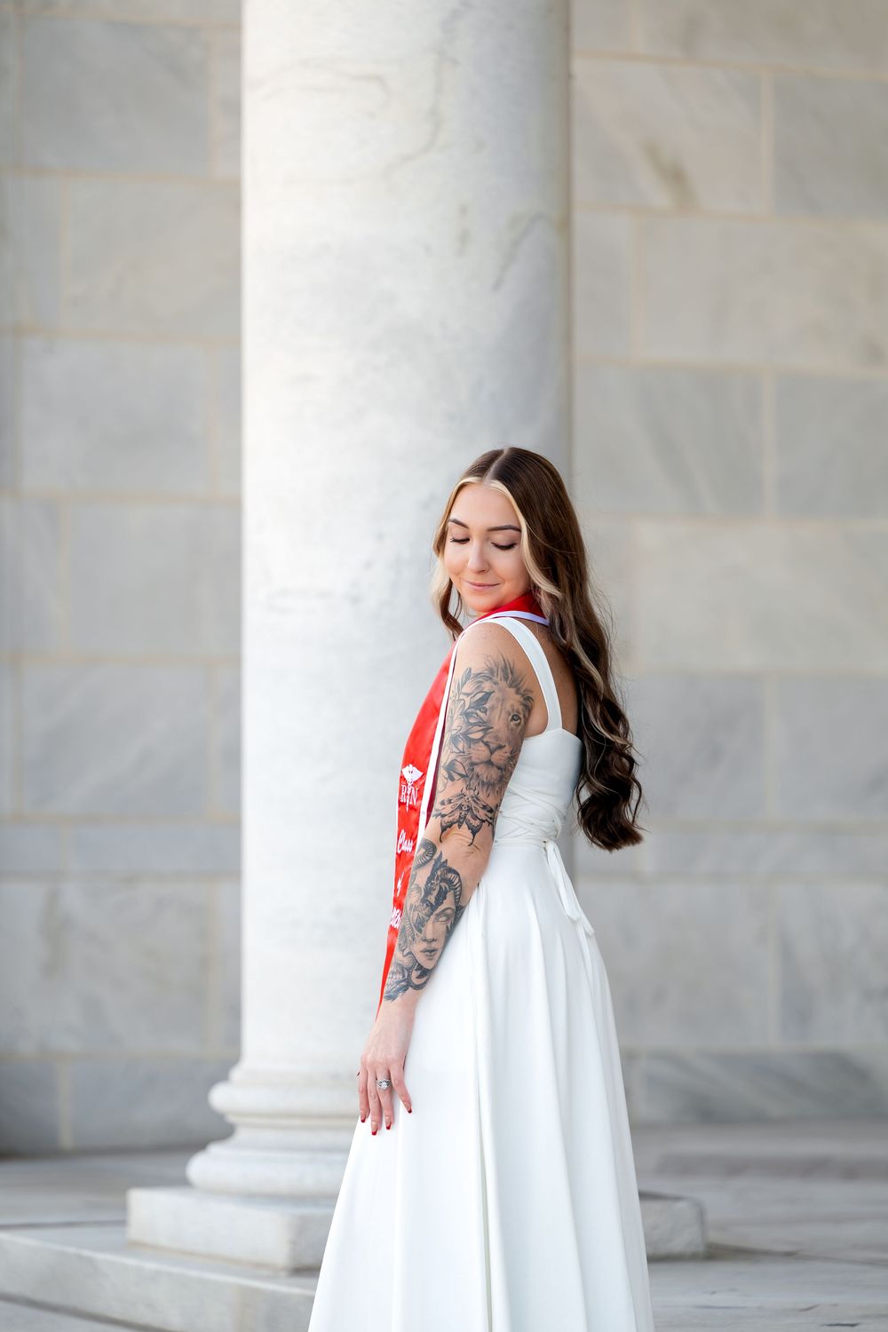 Young woman in white dress with red graduation sash and tattoos posing by marble columns.