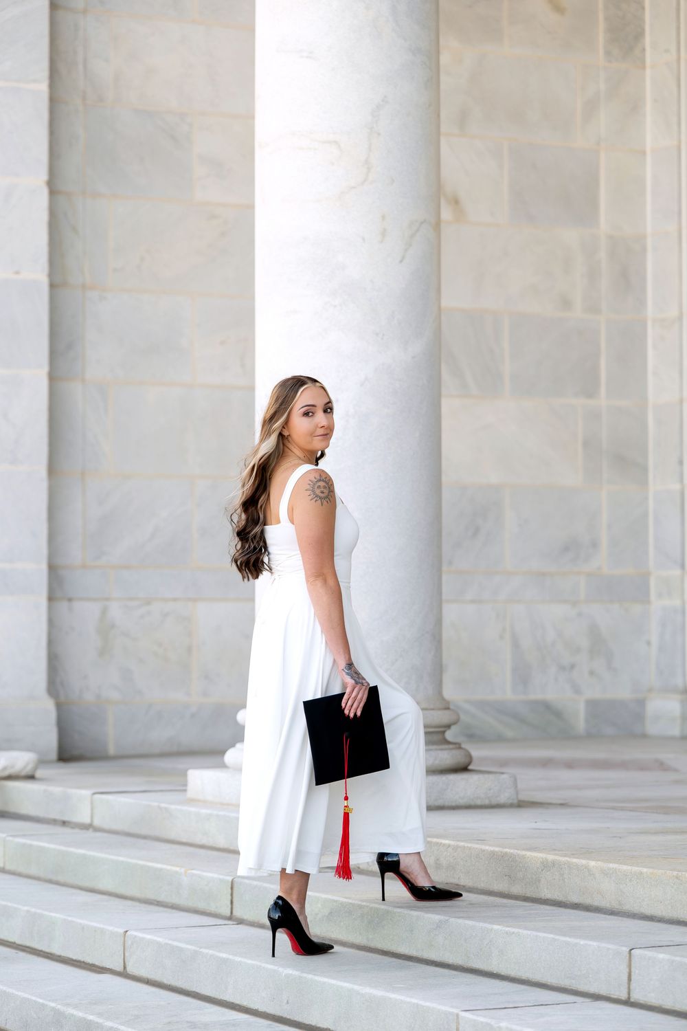 Young woman in white jumpsuit holding graduation cap with red tassel, posing on marble steps between columns.