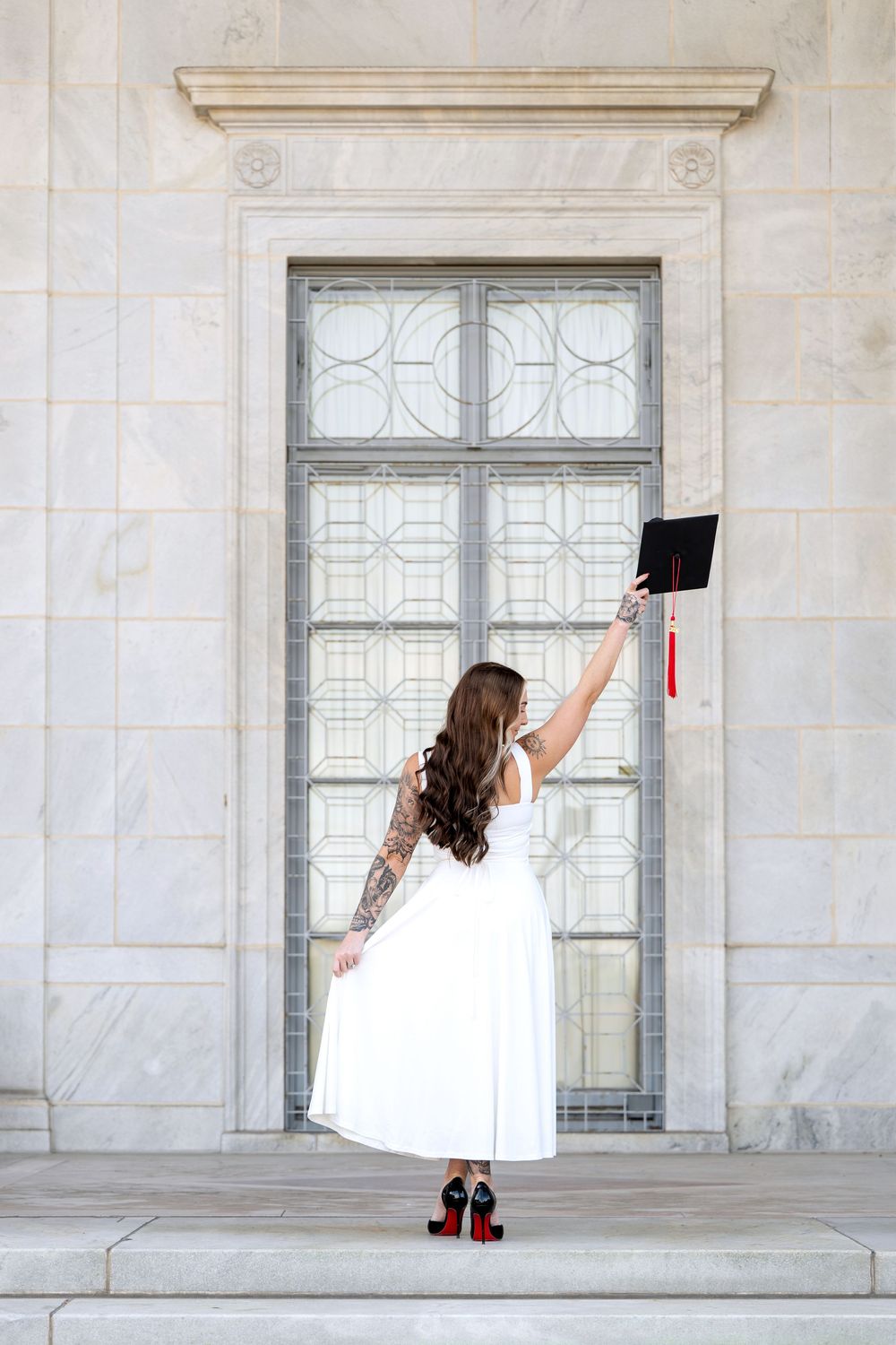 Woman in white dress and red heels raises graduation cap triumphantly on marble building steps.