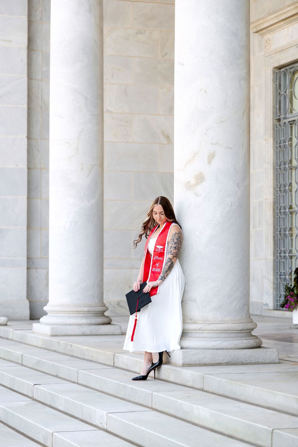 Graduate in white dress and red sash holds cap on marble steps between tall columns.