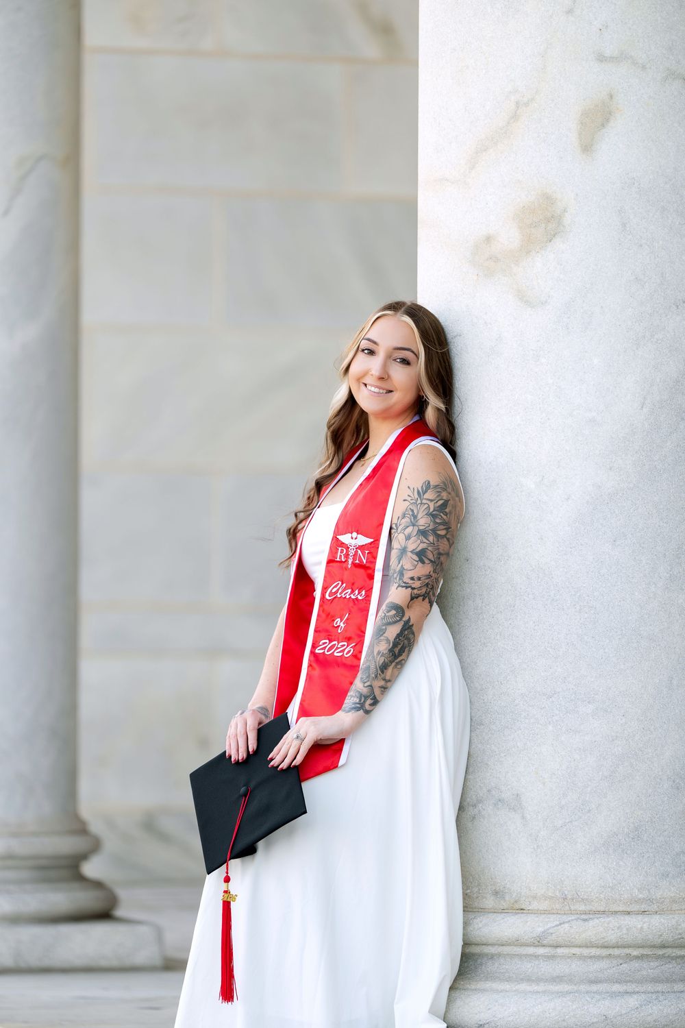 Smiling nursing graduate in white dress and red RN stole holds cap beside marble columns.