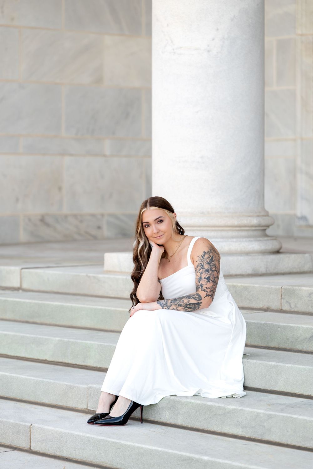 Woman in white dress with sleeve tattoos sits on marble steps beside columns, wearing black heels.