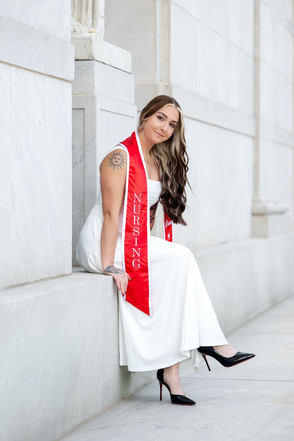Nursing graduate in white dress and red sash poses confidently in black heels against marble columns.