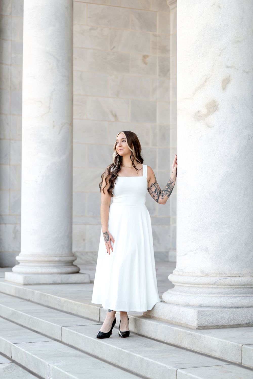 Elegant woman in white midi dress with tattoed arm poses between marble columns on stone steps.