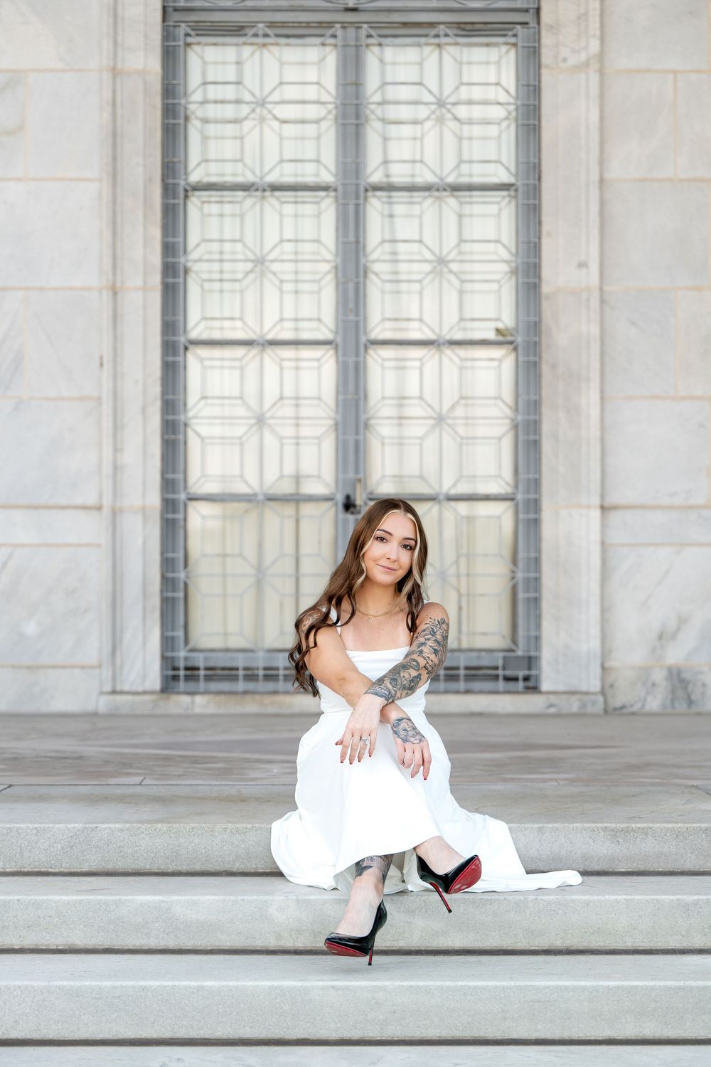 Woman in white dress and red-sole heels sits gracefully on stone steps before ornate building doors.