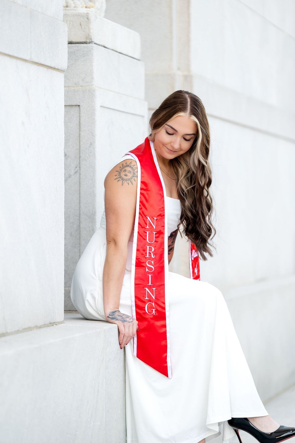 Nursing graduate in white dress with red sash sits on marble steps, celebrating her achievement.