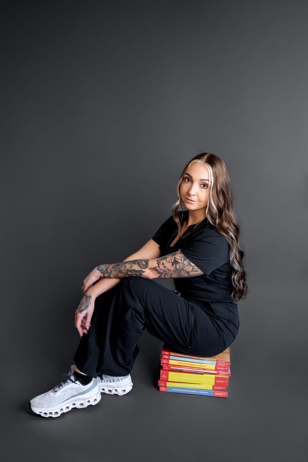 Woman in black outfit with tattoos sits on stacked books against gray background.