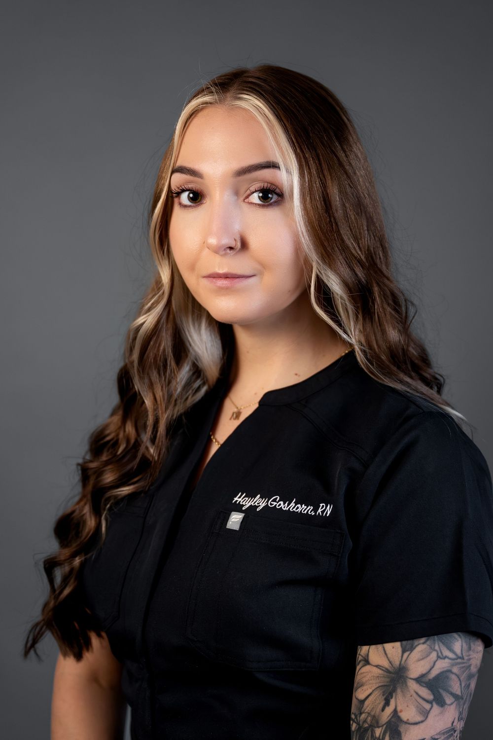 Professional headshot of Hayley Goshorn RN in black scrubs with floral arm tattoo on gray background.