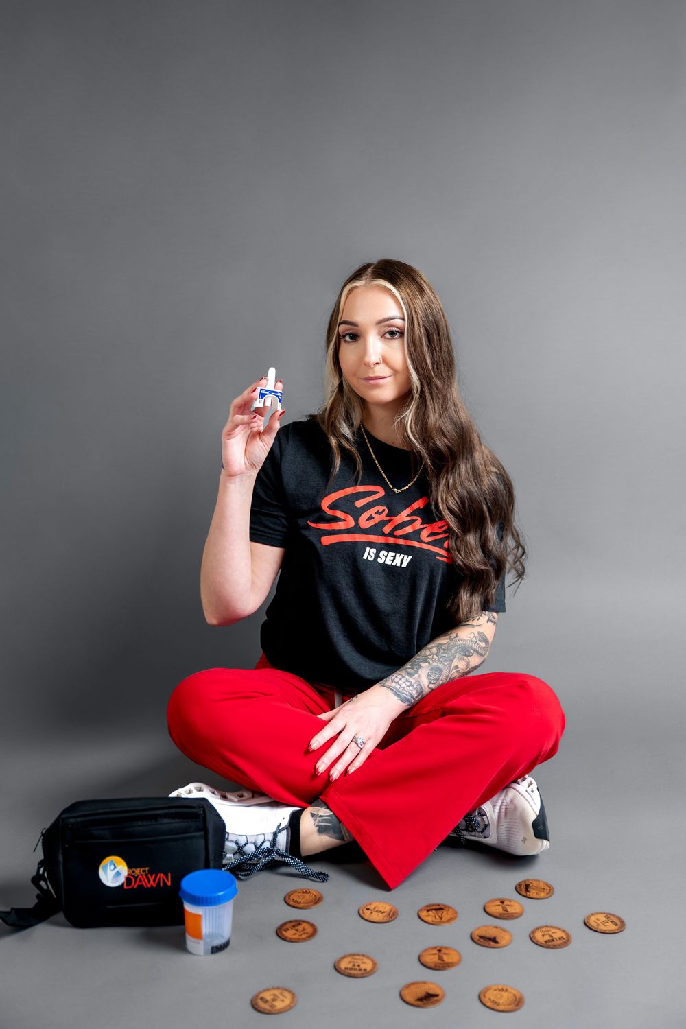 Woman in Sobe Is Sexy t-shirt sitting cross-legged holding nasal spray surrounded by coins and medical bag.