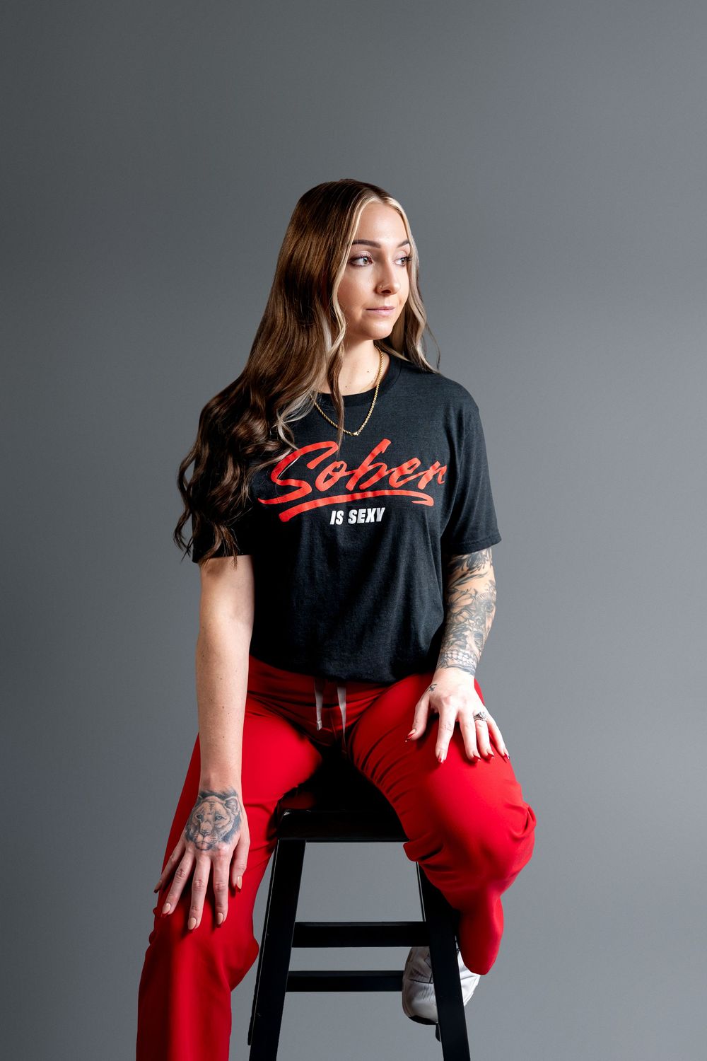 Young woman with long brown hair sits on stool wearing black Sober is Sexy tee, red pants, and tattoos on gray background.