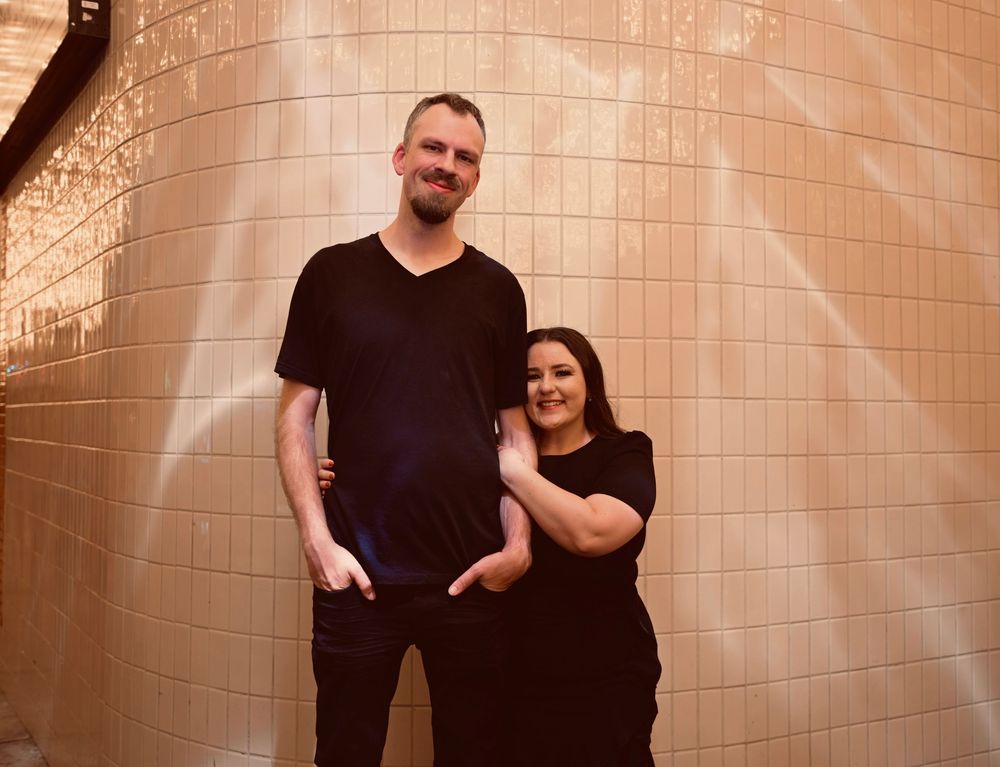 A tall man and a woman in black outfits pose smiling against a pink tiled wall.