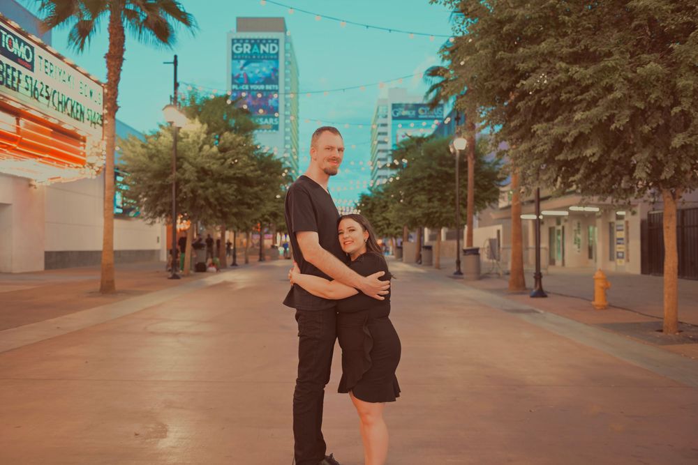 A couple poses on a Las Vegas street at dusk, the man embracing the pregnant woman from behind near the Fremont Street area.