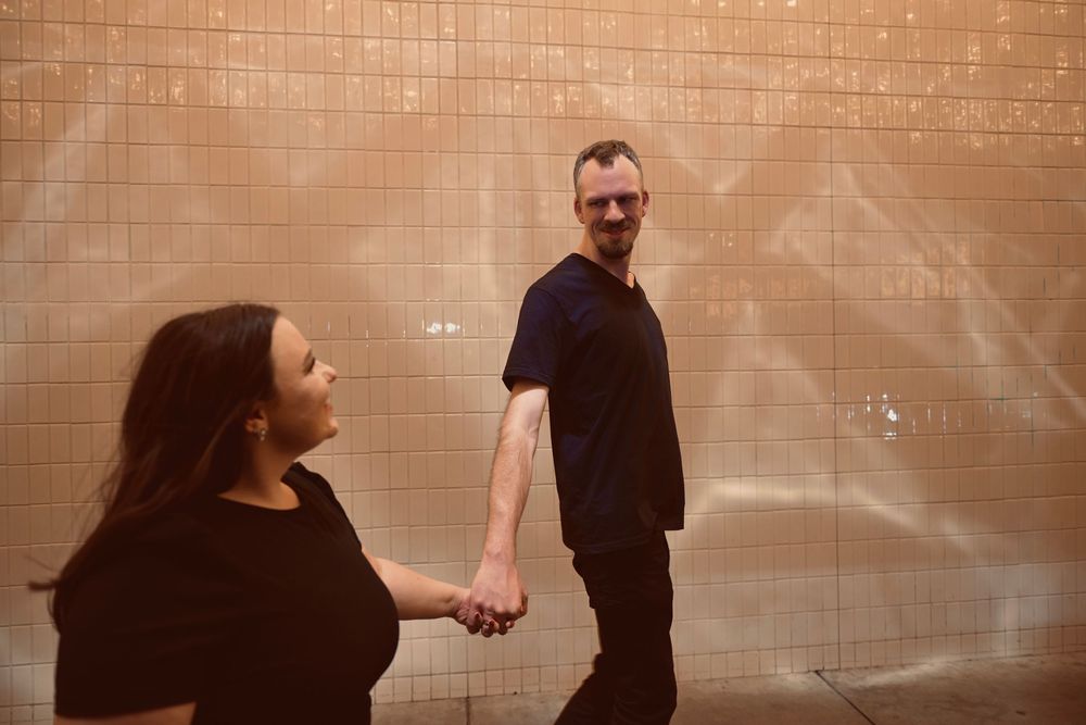 A couple holds hands in front of a glowing tiled wall, the man glancing back over his shoulder.