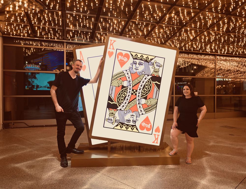 Two people posing with a giant King of Hearts playing card display under sparkling lights at a casino venue.