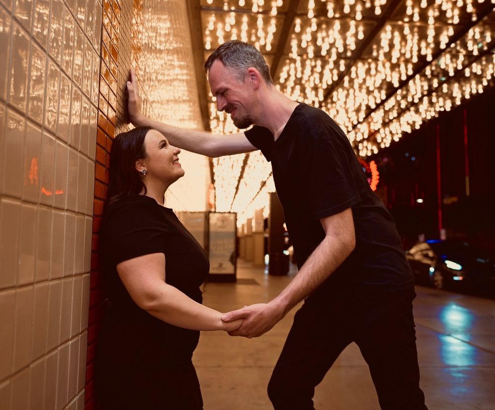 Couple in black outfits share a romantic moment under warm string lights in a dimly lit venue.