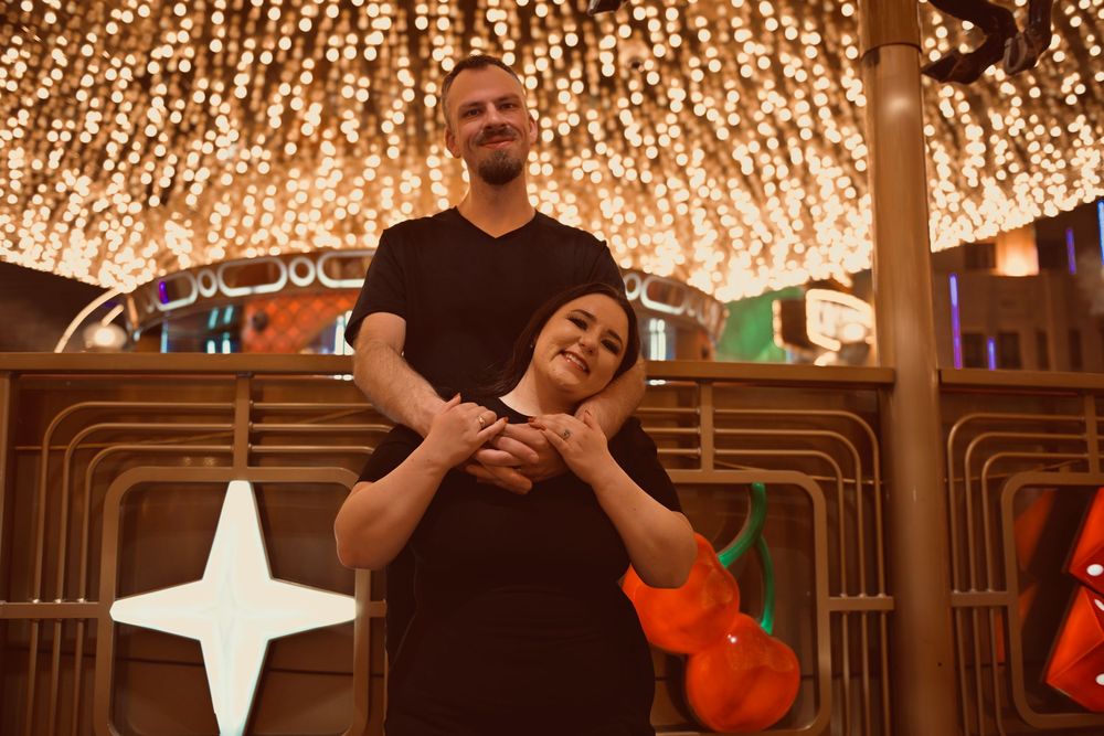Couple posing under glowing string lights at a festive venue, man standing behind woman with hands clasped together.