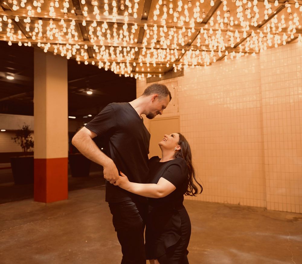 Couple in matching black outfits dancing romantically under warm string lights in an elegant venue.