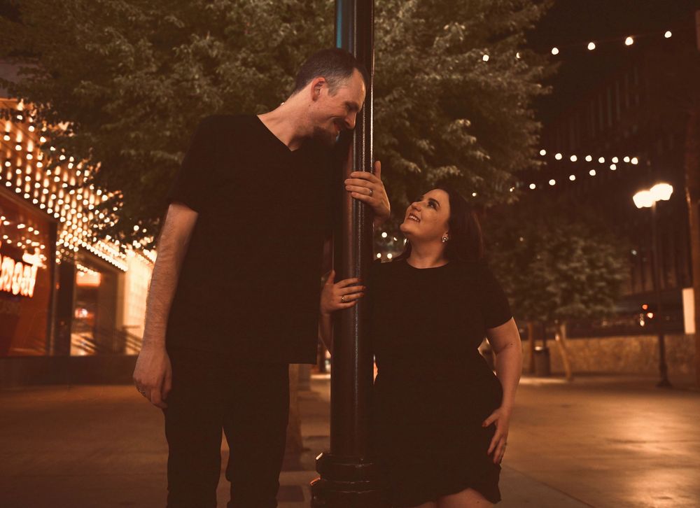 Couple sharing a romantic moment by a lamp post on a city street at night with string lights.