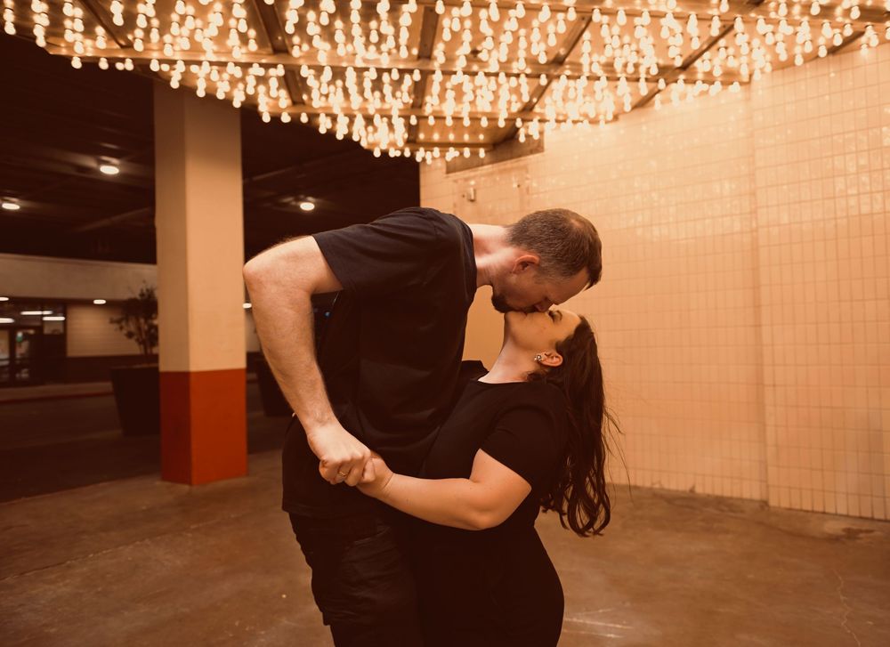 Romantic couple sharing a passionate kiss and dip under warm string lights in an indoor venue.