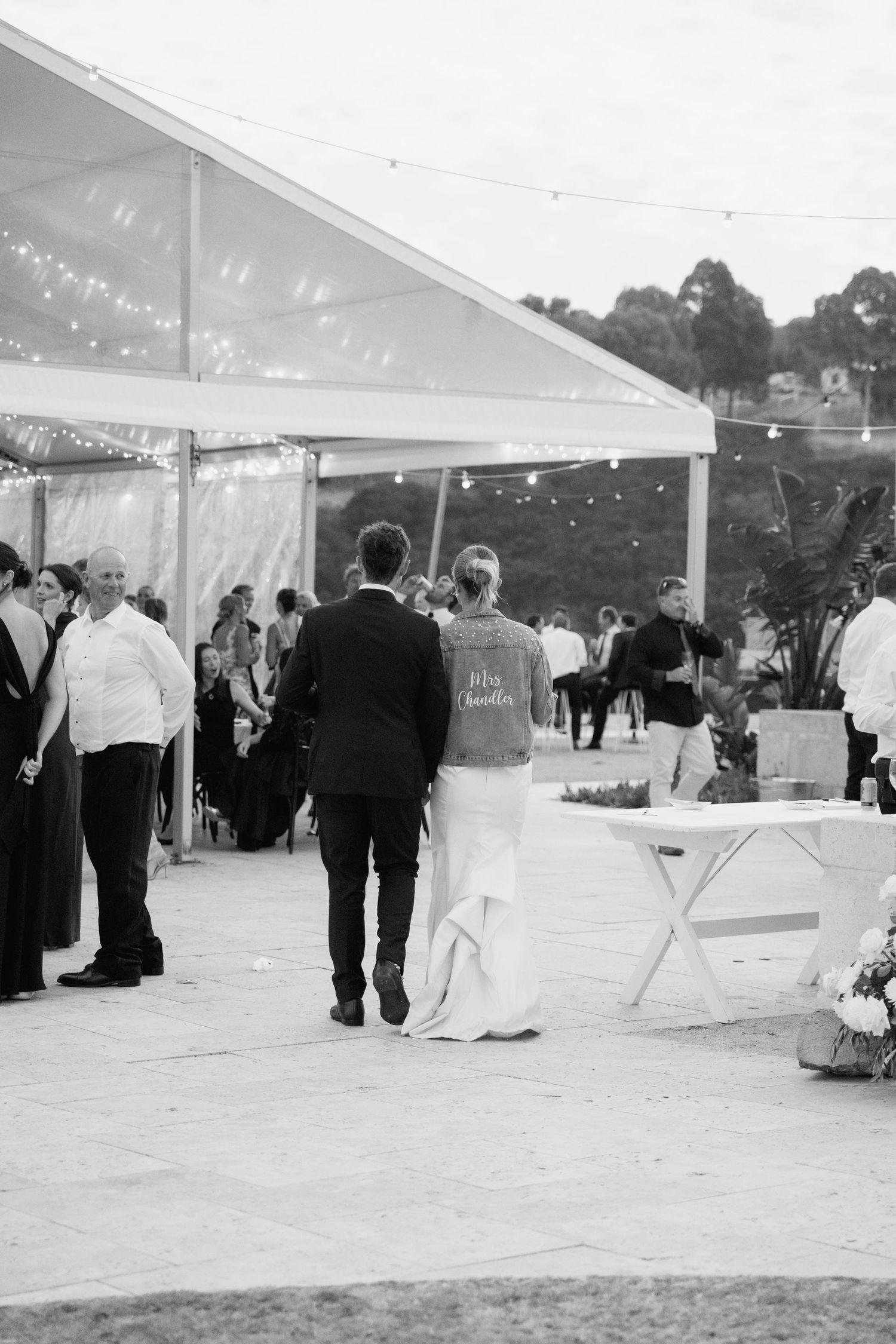 Black and white photo of bride and groom walking hand in hand at outdoor wedding reception under marquee with fairy lights.