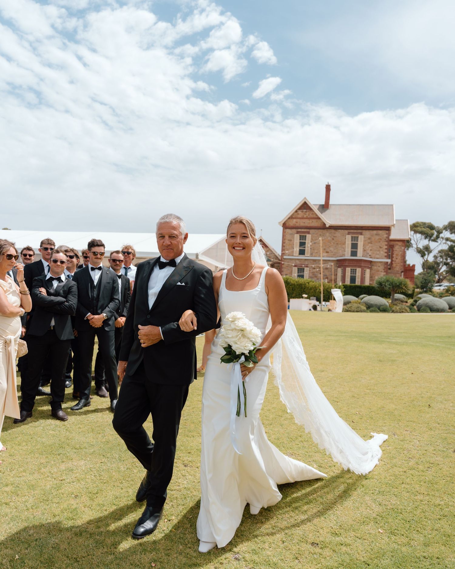 Bride in white gown walks down outdoor aisle with escort in tuxedo, guests watching, historic stone building backdrop.