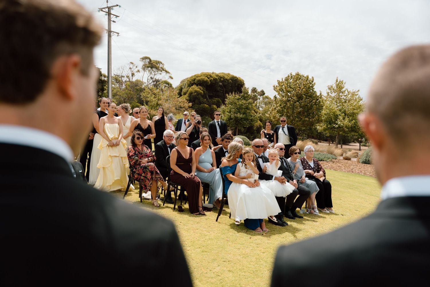 Outdoor wedding ceremony with guests seated on chairs on green lawn under bright sky.
