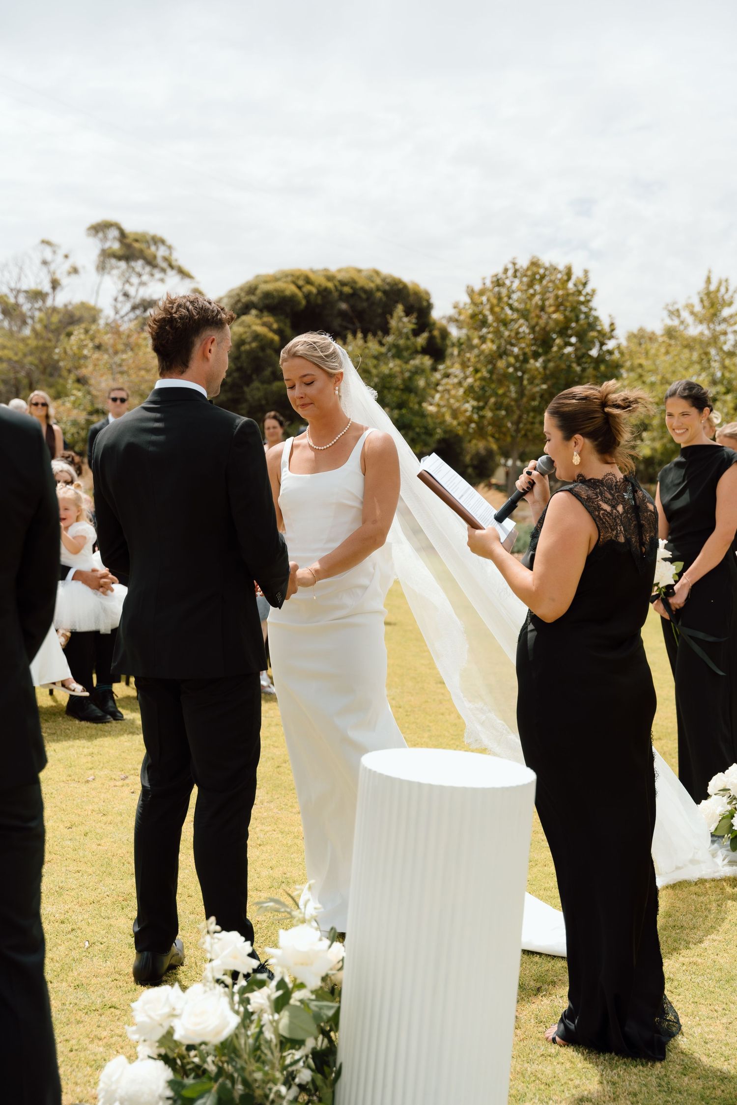 Bride and groom exchange vows at outdoor wedding ceremony while officiant holds veil in black dress.