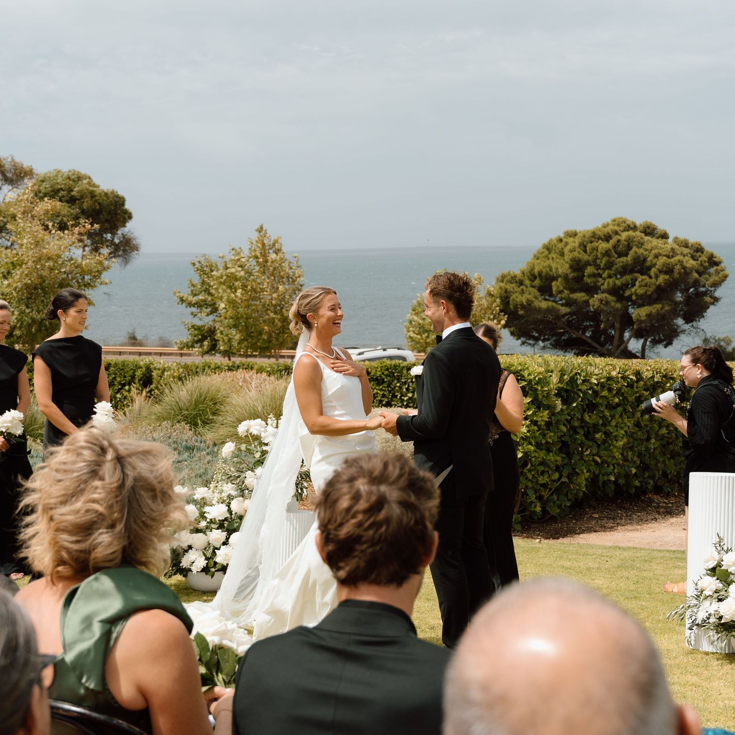 Bride and groom exchanging vows at an outdoor seaside wedding ceremony with white floral arrangements.
