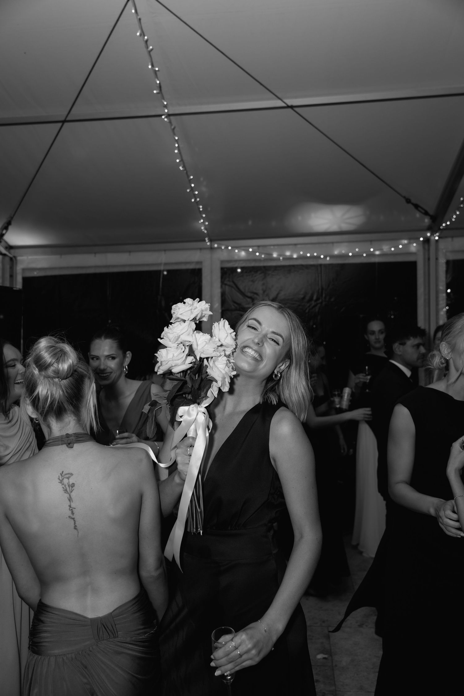 Woman in black dress laughing and holding bridal bouquet at wedding reception under string lights.