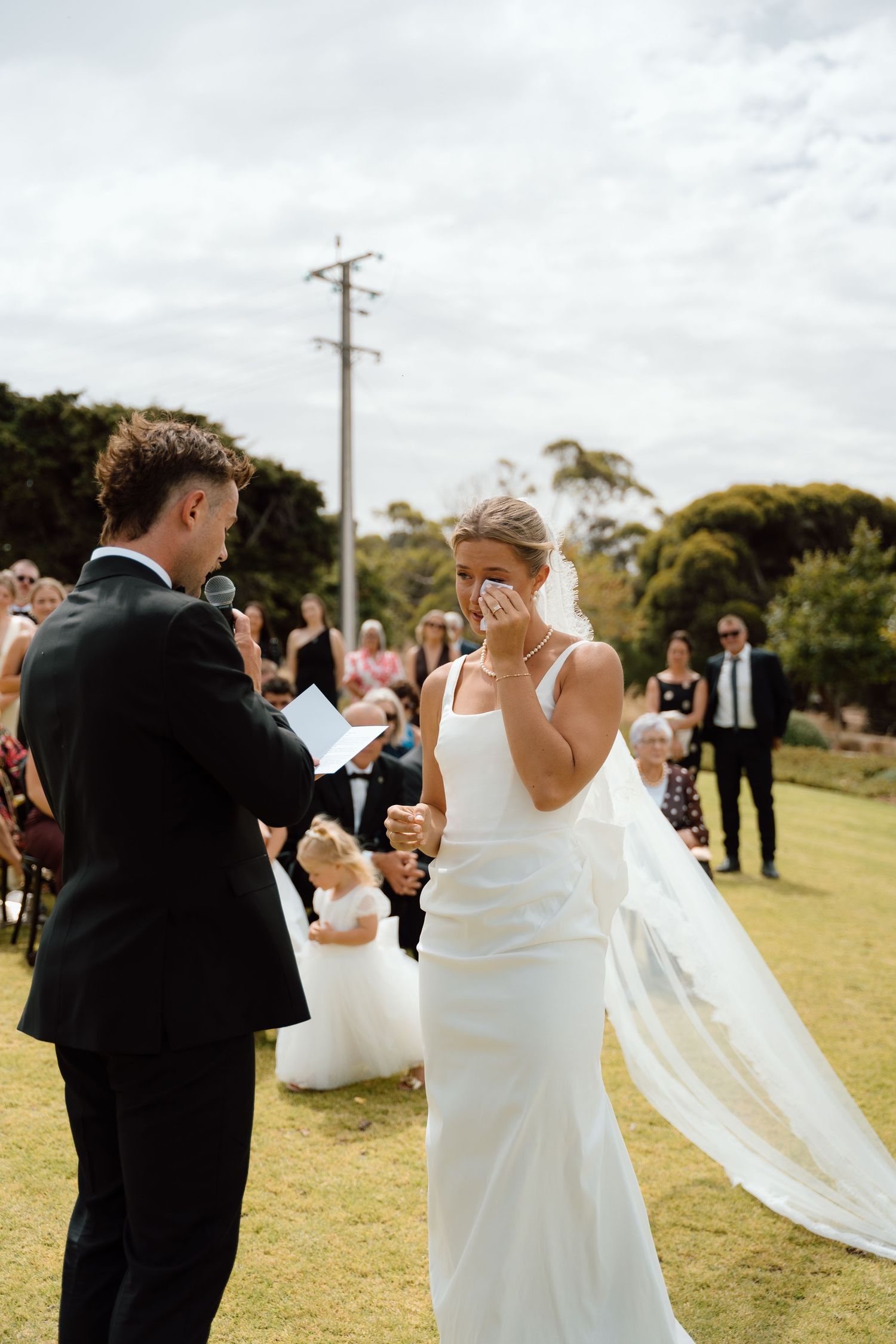Emotional bride in white gown wiping tears as groom reads vows at outdoor ceremony.