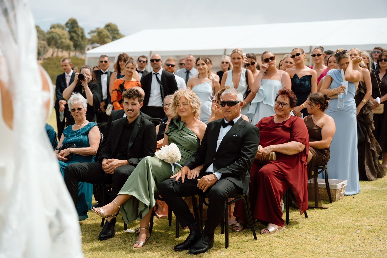 Outdoor wedding ceremony guests seated on chairs, wearing formal attire, smiling and watching the ceremony on a sunny day.