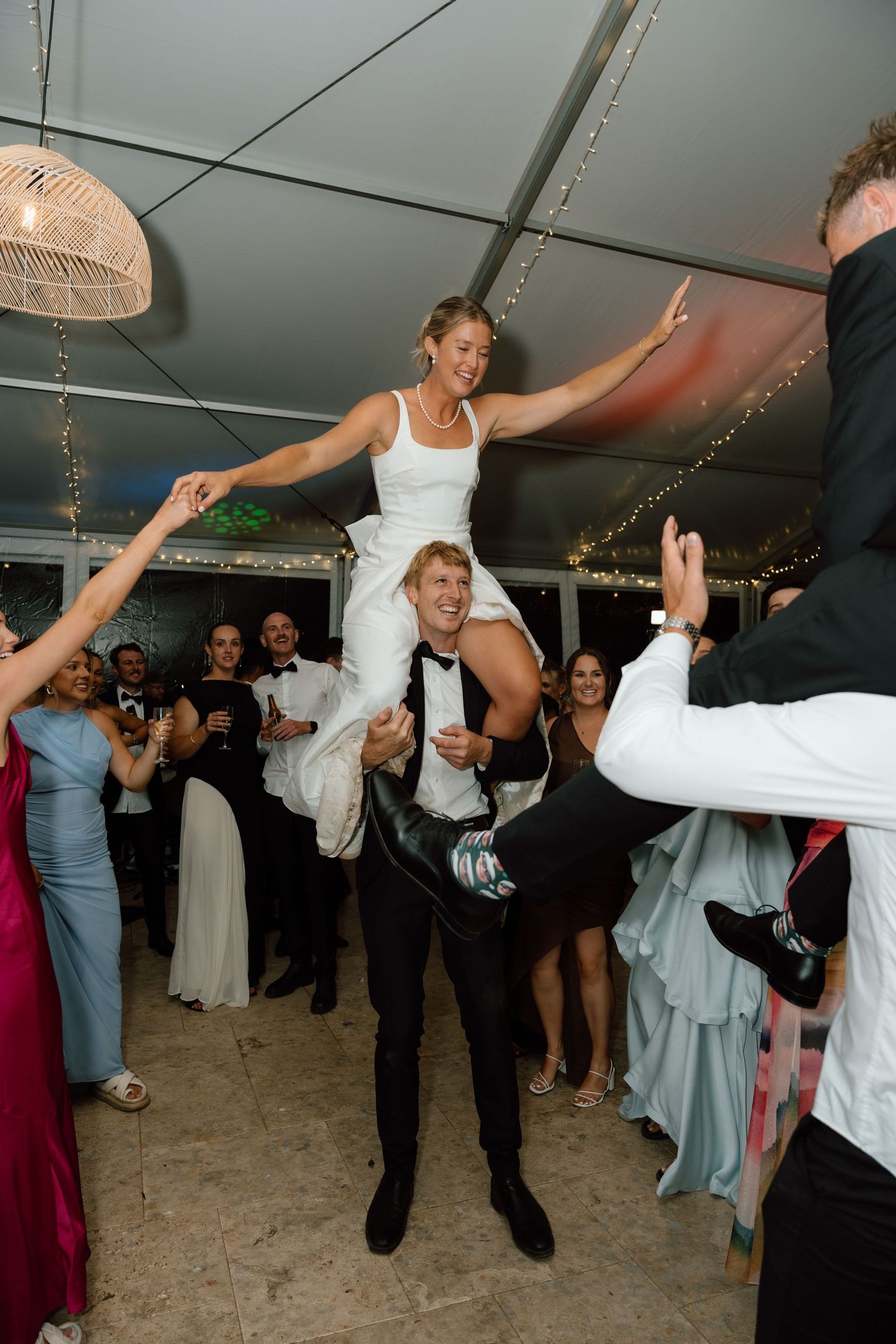 Bride lifted by groom on wedding dance floor surrounded by celebrating guests in elegant marquee reception.