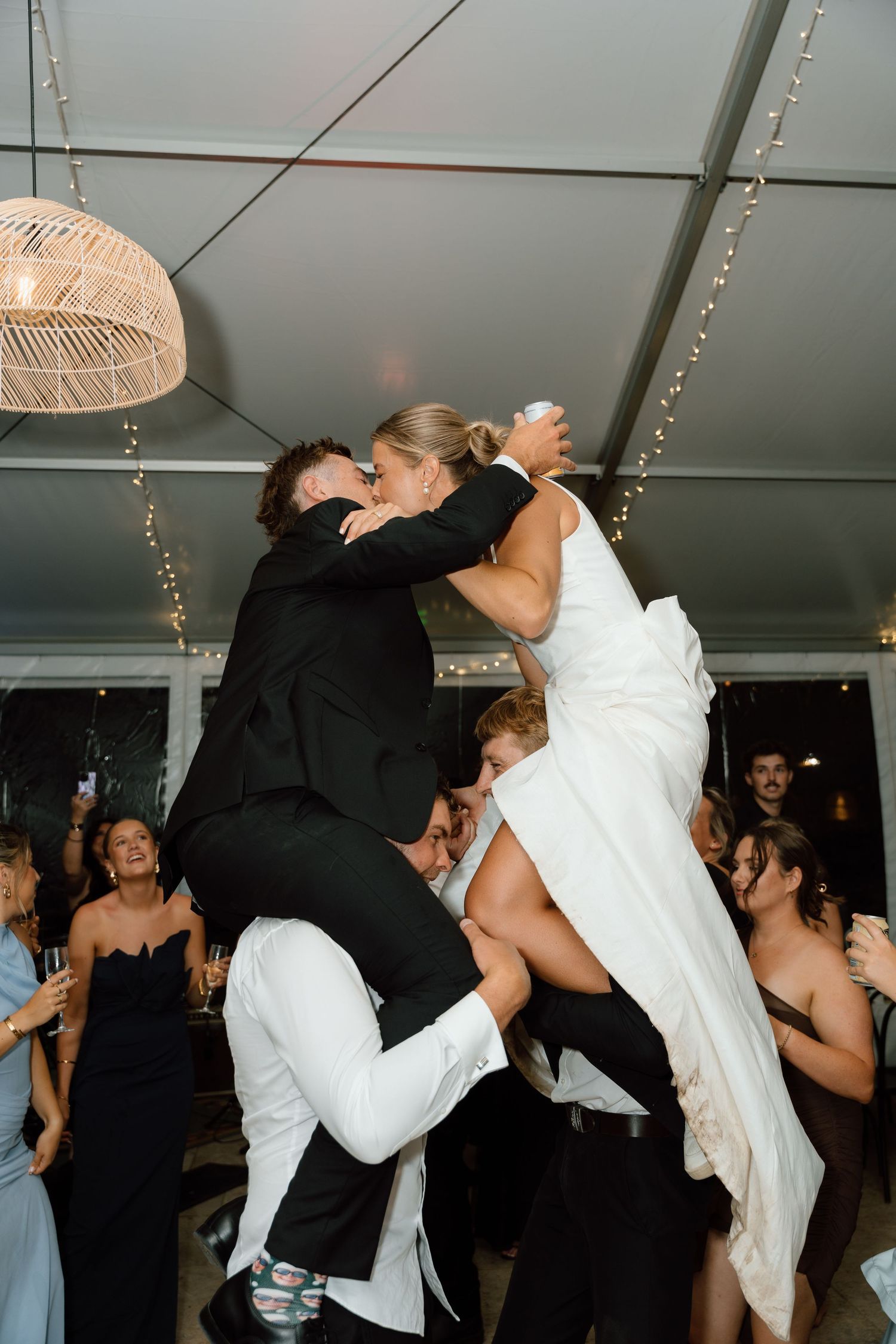 Bride and groom share a passionate kiss while being lifted by wedding guests on the dance floor.