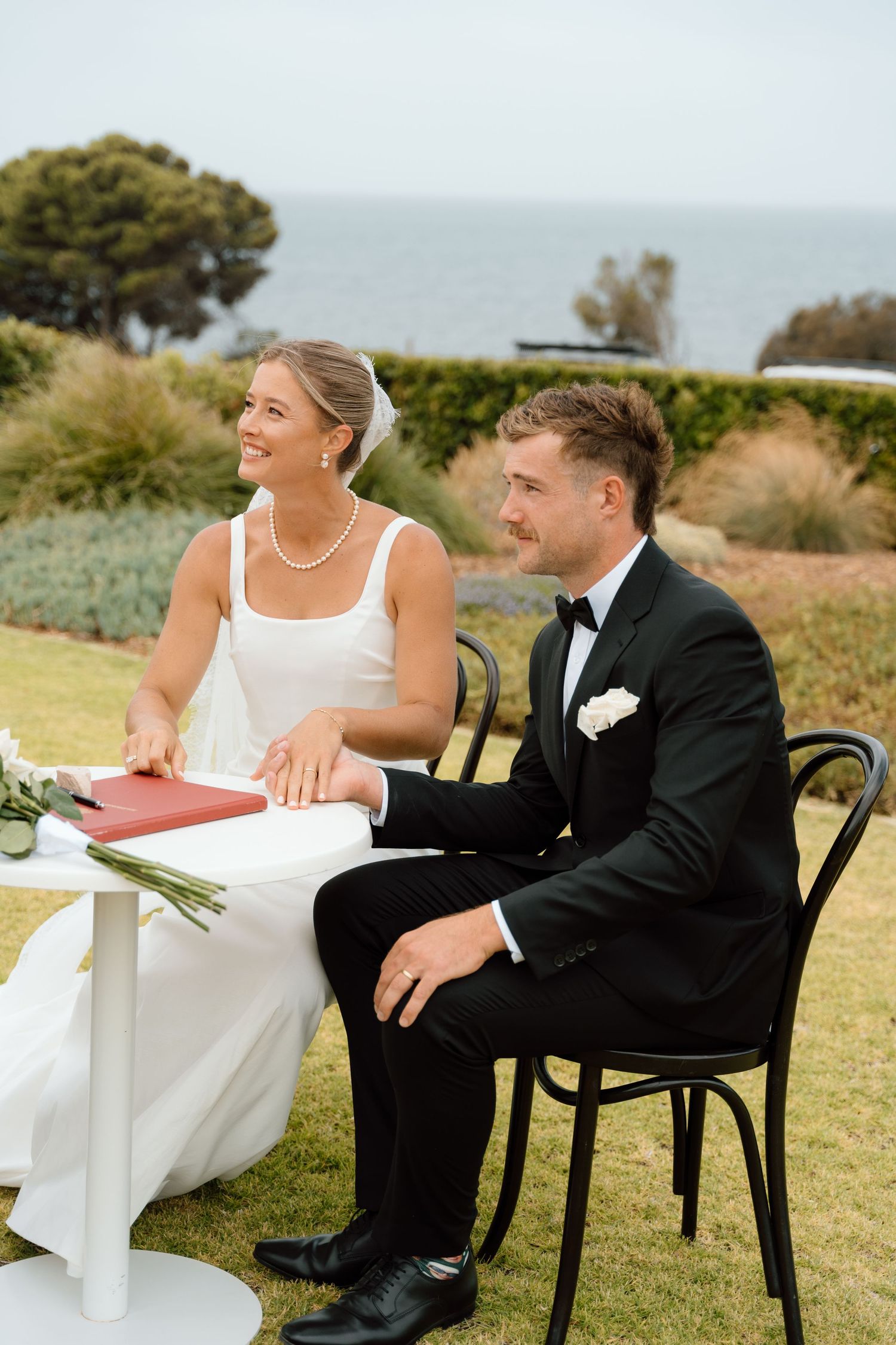 Bride in white gown and groom in black tuxedo sit at signing table during outdoor wedding ceremony with ocean backdrop.