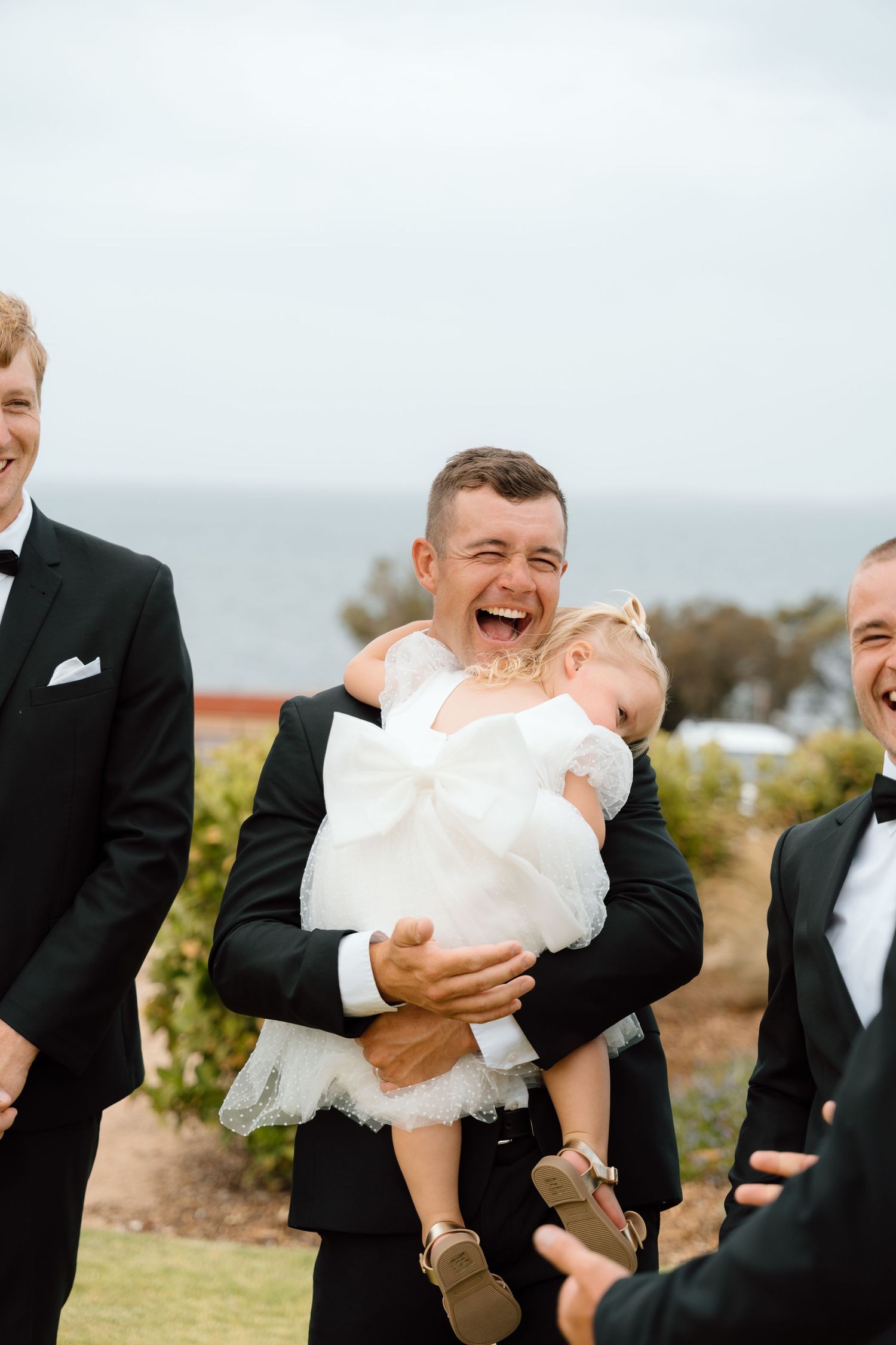 Laughing groomsman joyfully holds flower girl in white dress at outdoor coastal wedding ceremony.