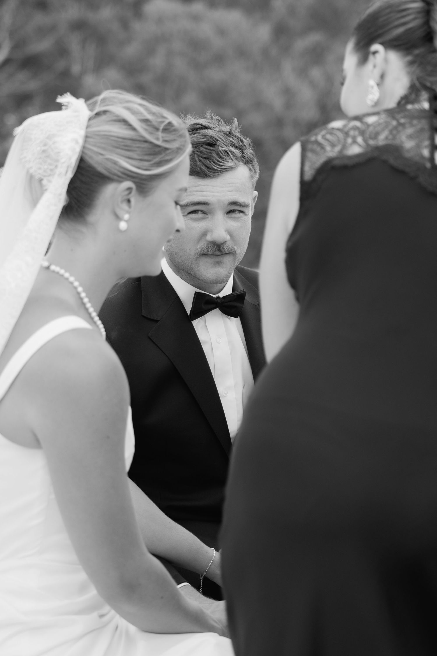 Emotional groom holds brides hands during outdoor wedding ceremony in black and white photo.