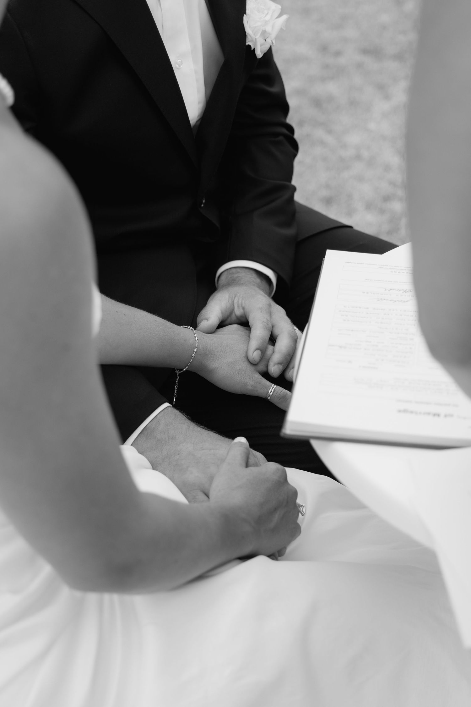 Black and white close-up of bride and groom holding hands during wedding signing ceremony.