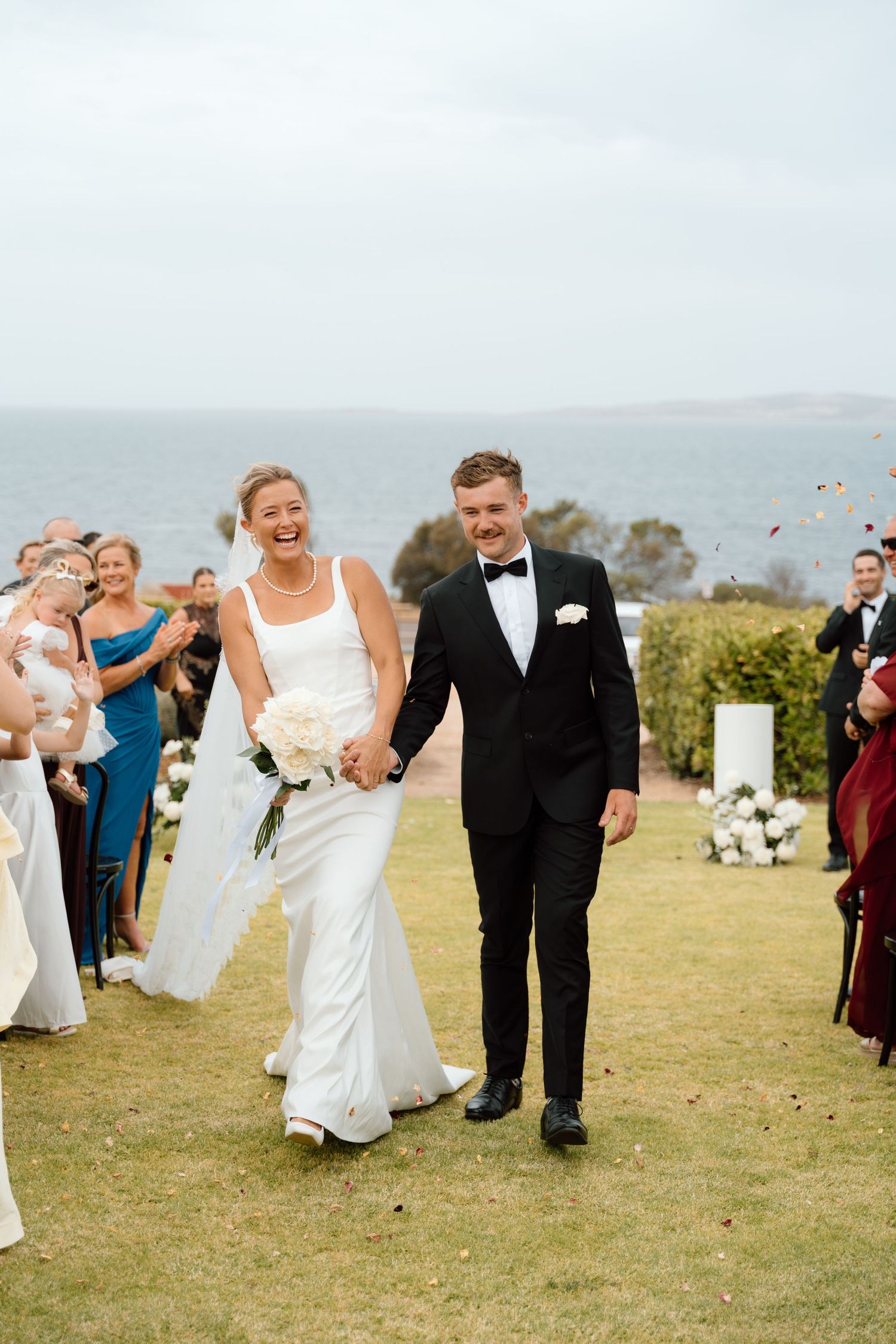 Bride and groom walk down outdoor aisle holding hands, laughing, with ocean backdrop and guests cheering.