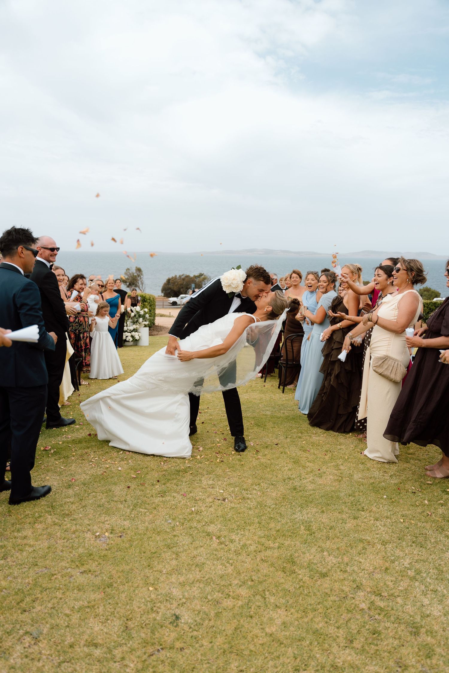 Bride and groom share romantic dip kiss as guests toss flower petals during outdoor wedding ceremony by the ocean.