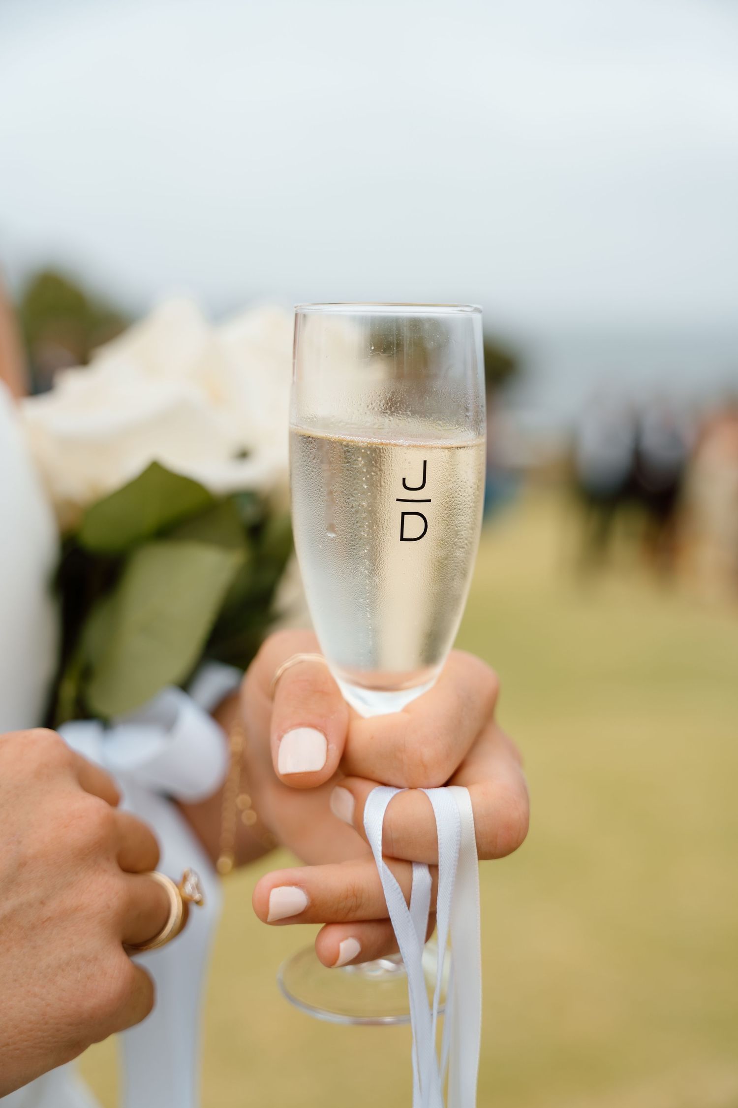 Hand holding a personalized champagne flute with initials at a wedding celebration outdoors.