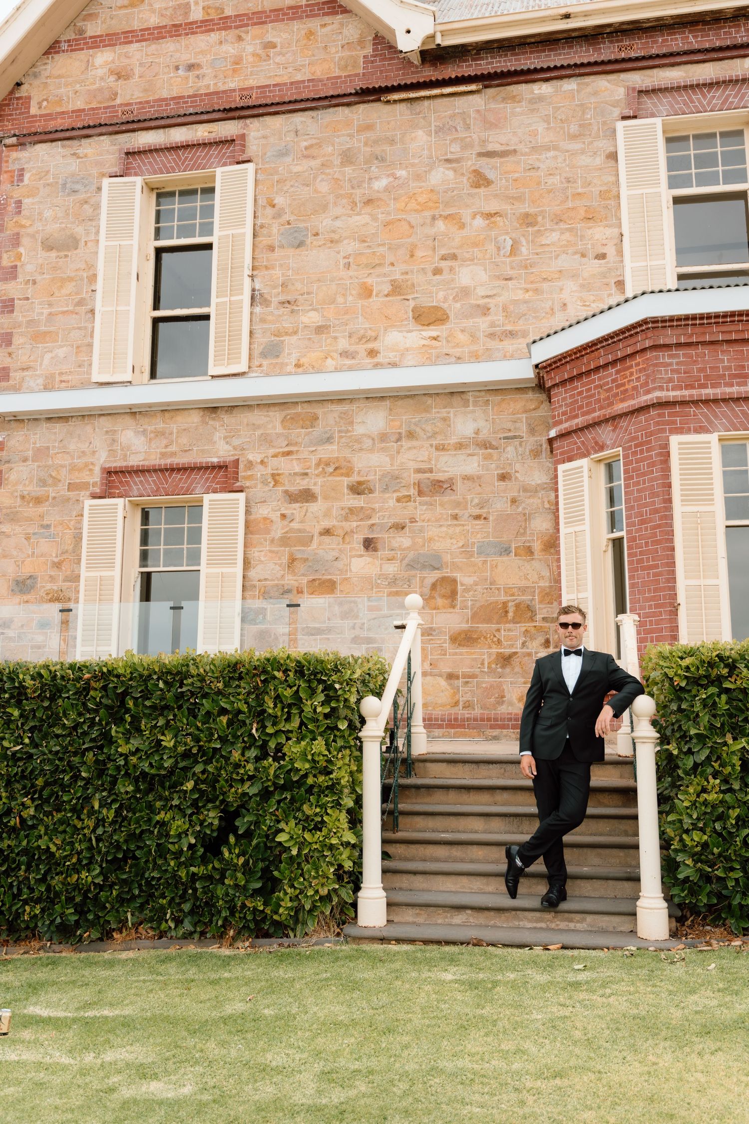 Dapper groom in black tuxedo and sunglasses poses on steps of elegant historic stone building before wedding ceremony.